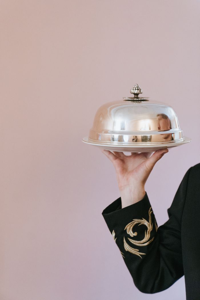 Close-up of a waiter holding a silver serving tray on a light pink background.